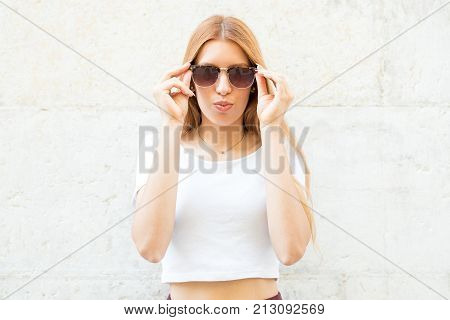 Fashionable girl in sunglasses puckering lips and looking at camera. Modern young woman putting sunglasses on. Style statement concept