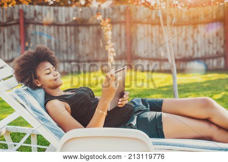 Attractive young Brazilian lady laying on lounge and chatting with friend via tablet pc; calm black teenage female is chilling on chaise-longue in park and watching music videos with digital pad