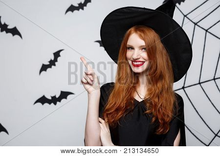 Halloween Witch Concept - Happy Halloween Witch Pointing Finger On Side Over Dark Grey Studio Backgr