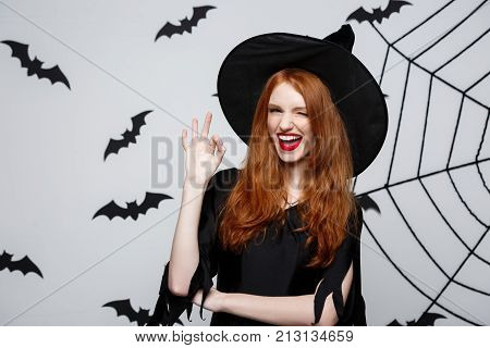 Halloween Witch Concept - Happy Halloween Ginger Hair Witch Holding Ok Sign With Fingers Posing Over
