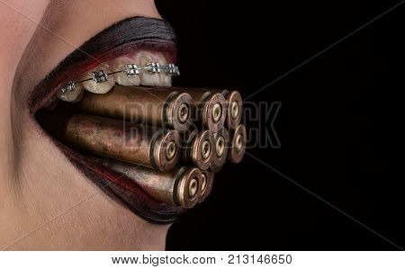Bullets from revolver or hand pistol between teeth with braces. Dentistry and orthodontics alignment of teeth as a weapon. Female mouth with bullets with black lipstick on black background