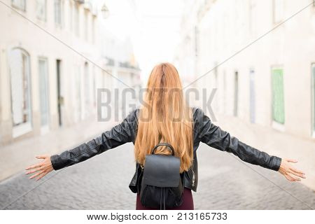 Redhead student girl enjoying freedom in city. Rear view of carefree woman with leather satchel wearing jacket outstretching arm opening her heart to new achievements. Vacation concept