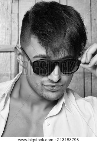 Sexy serious sensual muscular young macho man with bare torso in white shirt and sun glasses standing indoor on wooden background vertical picture