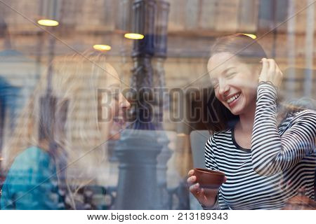 Two student girls smiling laughing while relaxing at university canteen during lunch break. Shot of attractive cheerful ladies talking drinking tea through big window of modern cafe or restaurant.