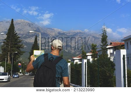 a man stands on the road in the village and looks at the tourist map dressed in a T-shirt and cap. a man with a backpack stands on a background of high mountains.