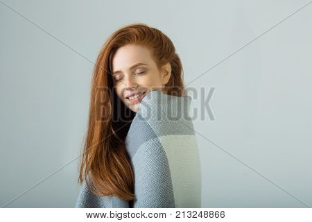 beautiful young girl with red hair warms up in a warm blanket on a light background