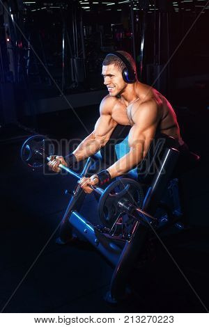Bodybuilding concept. Muscular man doing exercises for biceps with barbell on Scott's bench in gym and listening music on headphones. Bodybuilder lifting weights. Power fitness man training muscles
