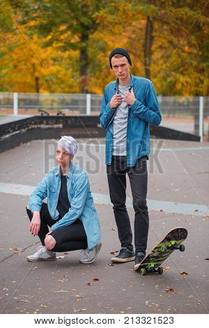 Young guy and girl skateboarders, outdoors on a bright autumn day, the guy's leg holds a skate, hands holding a shirt, the girl crouched.