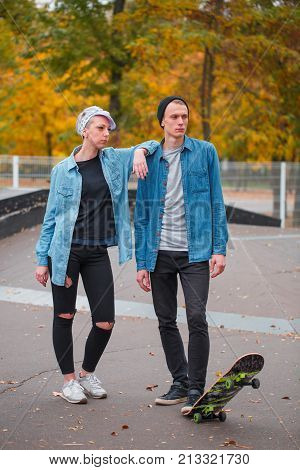 Young guy and girl skateboarders, outdoors on a bright autumn day at the foot of a guy holding a skate, the girl put her hand on the guy. They look aside.