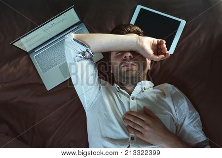 Tired caucasian young man in white shirt sleeping and keeping one hand above head, lying on bed near laptop. He is tired of work problems.