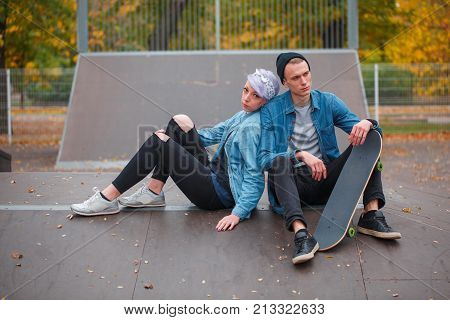 A young guy and a girl skateboarders, outdoors on a bright autumn day, sitting on a springboard at a man's skate lies between her legs, the girl rested her back on the guy's shoulder.