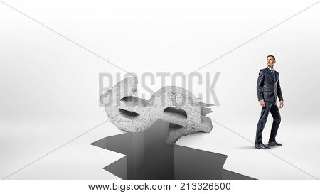 A businessman stands in half turn near a large earthquake crack on a white surface with a giant concrete dollar sign falling inside. Financial crisis. Recession and debt. Foreign exchange rates.