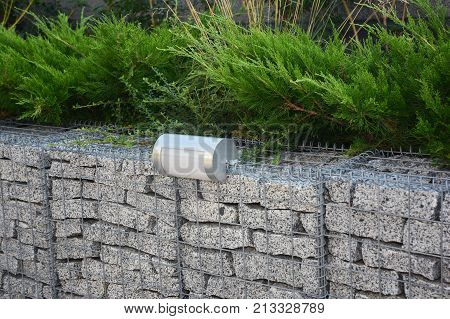 Gabion fence with stones in wire mesh and outdoor lighting. Gabion wire mesh fencing with natural stones and savin juniper.