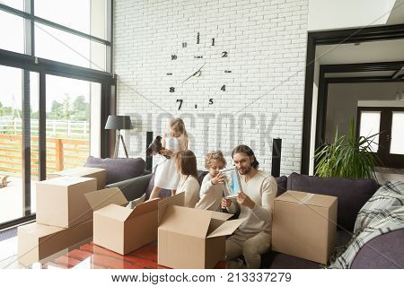 Young happy family with kids unpacking boxes together sitting on sofa in modern living room of country house moving settling in new home, children helping parents to pack stuff move out relocating