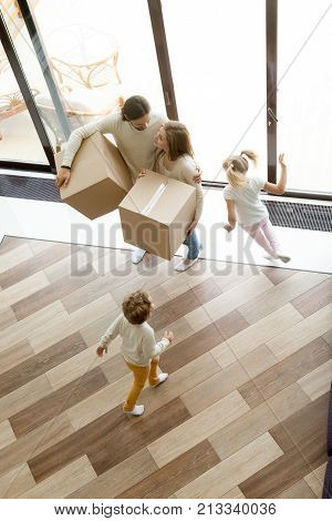 Family and children moving into new home, kids playing running around parents holding boxes standing in house hall together, happy couple with two children having fun move in, vertical top view