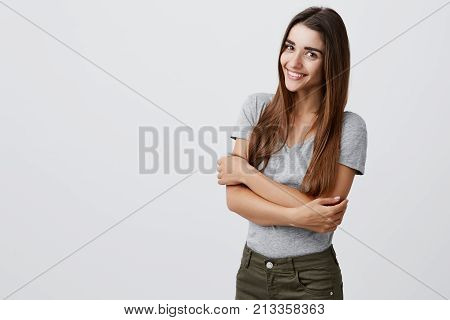 Cheerful young beautiful brunette caucasian student girl with long hair in casual stylish outfit smiling brightly, holding hands together, posing for university graduation photo in light studio. Copy space