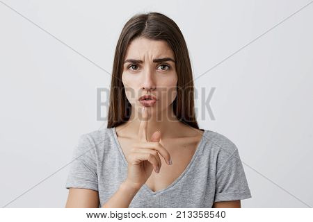 Close up portrait of young handsome sexy brunette caucasian woman with long hair in gray t-shirt holding index finger in front of lips, making hush gesture. Girl asking little kid to talk quieter in librar