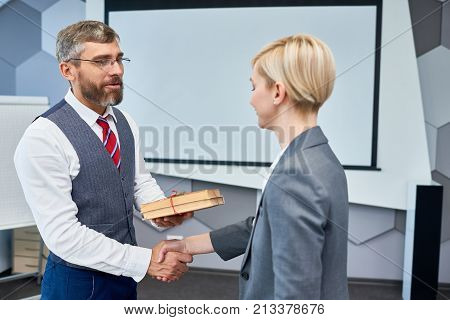 Portrait of bearded businessman shaking hands with female colleague presenting her gift for Boxing Day