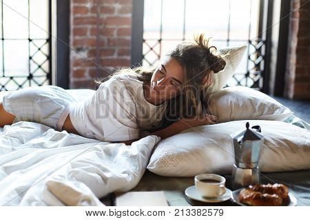 Cozy shot of gorgeous European female with unbrushed hair enjoying early hours in bed wearing stylish pajamas lying on white sheets and pillows with coffee cup pot and croissant on table