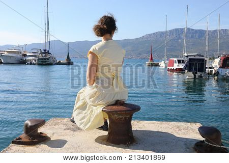 A woman in a yellow dress is sitting on a pier near yachts