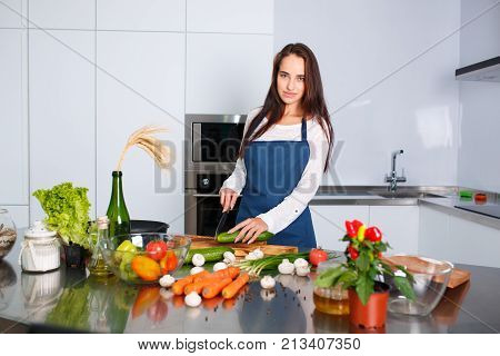 A young brunette in a blue apron with her hair in the kitchen looking at the camera and cutting a cucumber. On the table are scattered vegetables, green onions, pepper peas, champignons.