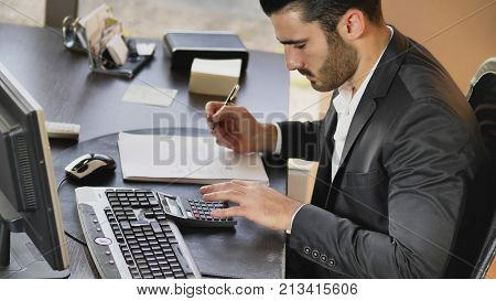 Confident man in formal wear working as accountant at desktop taking notes while using calculating machine.