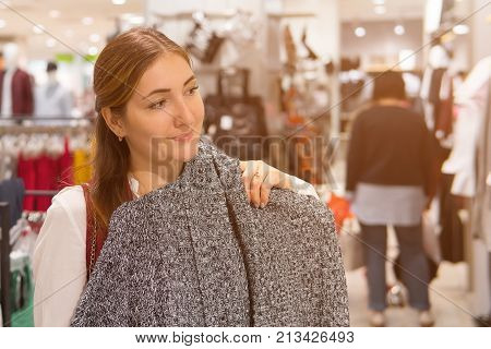 Young smiling woman trying on gray woolen jacket in clothing shop. Girl choosing warm clothes in fashion store