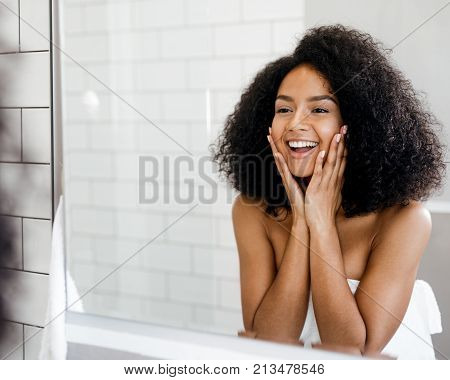 Happy woman admiring her skin in front of the bathroom mirror, touching face with hands