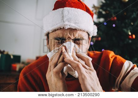 Close up of a sick man sitting on the floor after being woken up by nasal infection after Christmas eve's celebration. So he decided to use a tissue for sneezing. He hopes he will get better soon.