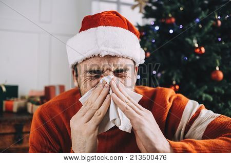 Another close up of a young man suffering from nasal infection. He is sneezing and using white tissue for that. It's not a good pleasure to be sick during Christmas holidays.