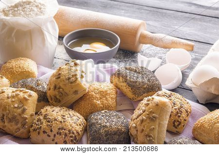 Freshly baked bread rolls and ingredients - Baking theme image with homemade buns decorated with poppy sesame flax and sunflower seeds and the ingredients for their making in the background.