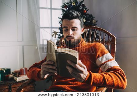 Another picture of good-looking guy sitting in the rocking chair at the Christmas tree and reading interesting book. He has almost finished reading it. Man decided to read a book while he is waiting for his friends to come to celebrate Christmas.