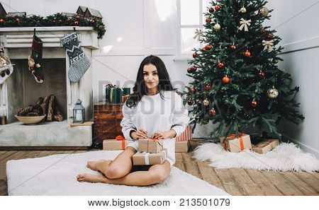 Awesome and stunning girl is sitting on the white furry carpet in a bright room. There are both fire place and Christmas tree behind her. She wears a white sweater and holding two boxes of presents for her friends. Young lady looks happy.
