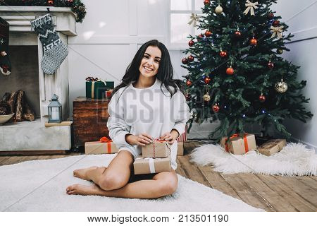 Another picture of attractive girl sitting on the white cozy carpet and smiling to the camera. She holds presents for her family on her knees and waiting for them in the room. Behind her there are a fireplace and a Christmas tree.