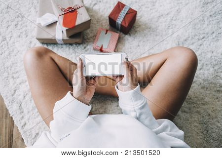 Close up of a girl sitting on the white carpet with her legs crossed and holding a phone with her hands. The phone's screen is white. Also there are some small boxes of Christmas gifts on the carpet. Cut view