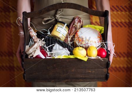 Female Hands Holding Basket With Colorful Eggs, Cake, Red Wine, Hamon Or Jerky And Dry Smoked Sausag