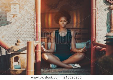 Young charming African American female with curly hair sitting inside of oriental pagoda on a matting and meditating halo appeared around her head she is feeling peaceful and calm reached nirvana