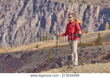 Tall backpacker with beard and sunglasses hold poles in hand. Sunny spring daybreak in rocky mountains. Hiker with backpack stand on rocky view point above valley.