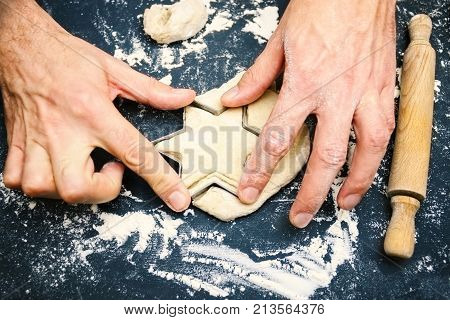 A photo of father's hands cutting the star with a cookie cutter on the wooden table. An overhead photo of man's hands flour wheat dough cookie cutter rolling pin on the rustic wooden table