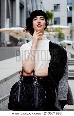 Stylish young female office worker in classic outfit in the city center wearing white turtle neck black beret and cardigan. Business city coffee break.