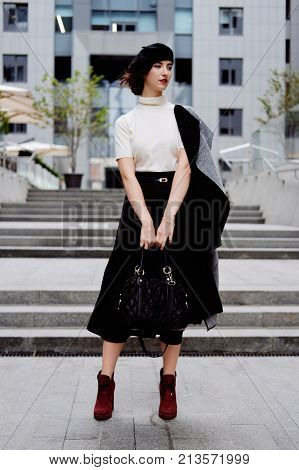 Stylish young female office worker in classic outfit in the city center wearing white turtle neck black beret and cardigan. Business city coffee break.
