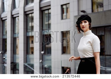 Stylish young female office worker in classic outfit in the city center wearing white turtle neck black beret and cardigan. Business city coffee break.