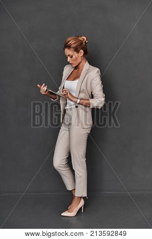 Young and smart. Full length of confident young woman in smart casual wear working on digital tablet while standing against grey background