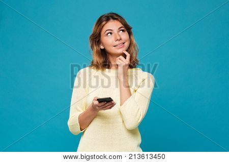 Pensive smiling woman in sweater holding smartphone and lookin up over blue background