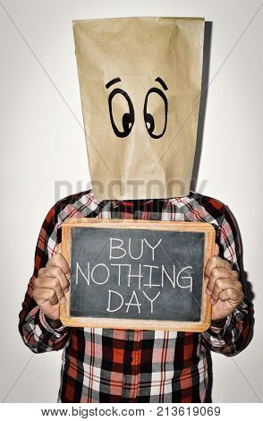 a young man wearing casual clothing with a paper bag in his head with a pair of eyes drawn in it shows a chalkboard with the text buy nothing day written in it