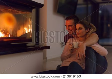 Young romantic couple sitting on the floor in front of fireplace,talking and drinking tea on cold winter night at home