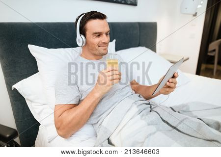 The invalid lies in bed and looks something on the tablet. He smiles. because he is happy. Next to his bed is his black wheelchair. It lies on a large white step.