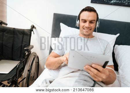 The invalid lies in bed and looks something on the tablet. He smiles. because he is happy. Next to his bed is his black wheelchair. It lies on a large white step.