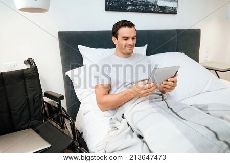 The invalid lies in bed and looks something on the tablet. He smiles. because he is happy. Next to his bed is his black wheelchair. It lies on a large white step.