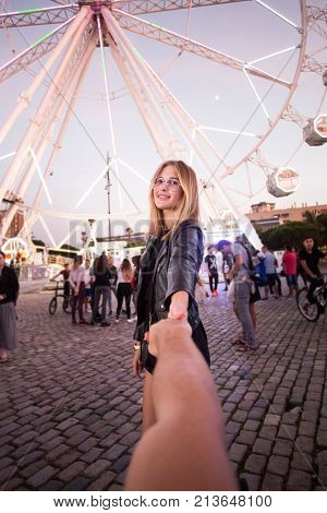 POV shot of boyfriend holding his girlfriend attractive young blonde woman by hand in front of ferris wheel at amusement park or carnival or festival enjoying romantic date together concept relationship goals at sunset